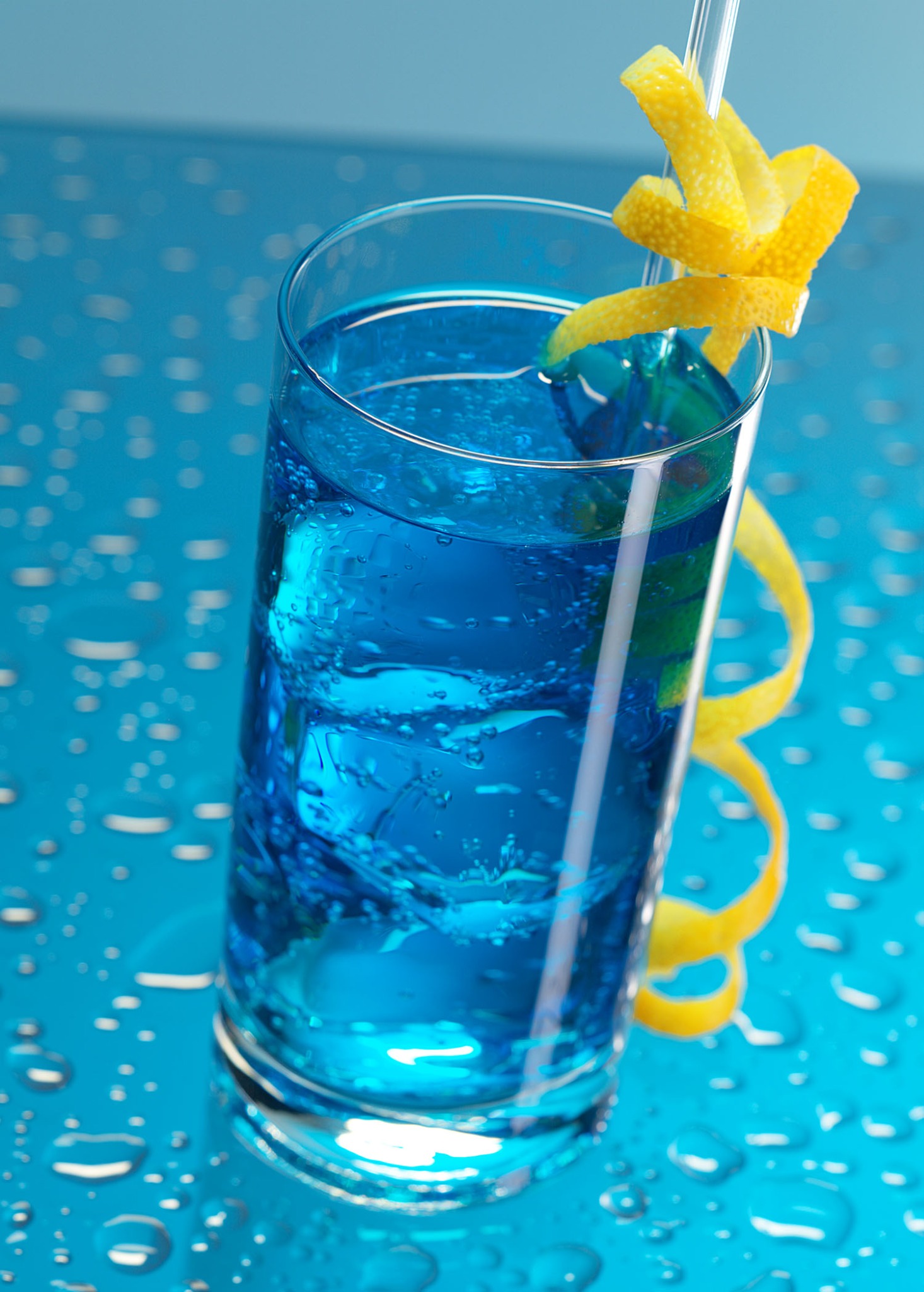 Szklanka z błękitnym napojem i lodem, ozdobiona spiralą ze skórki cytryny na tle niebieskiej powierzchni z kroplami wody.
