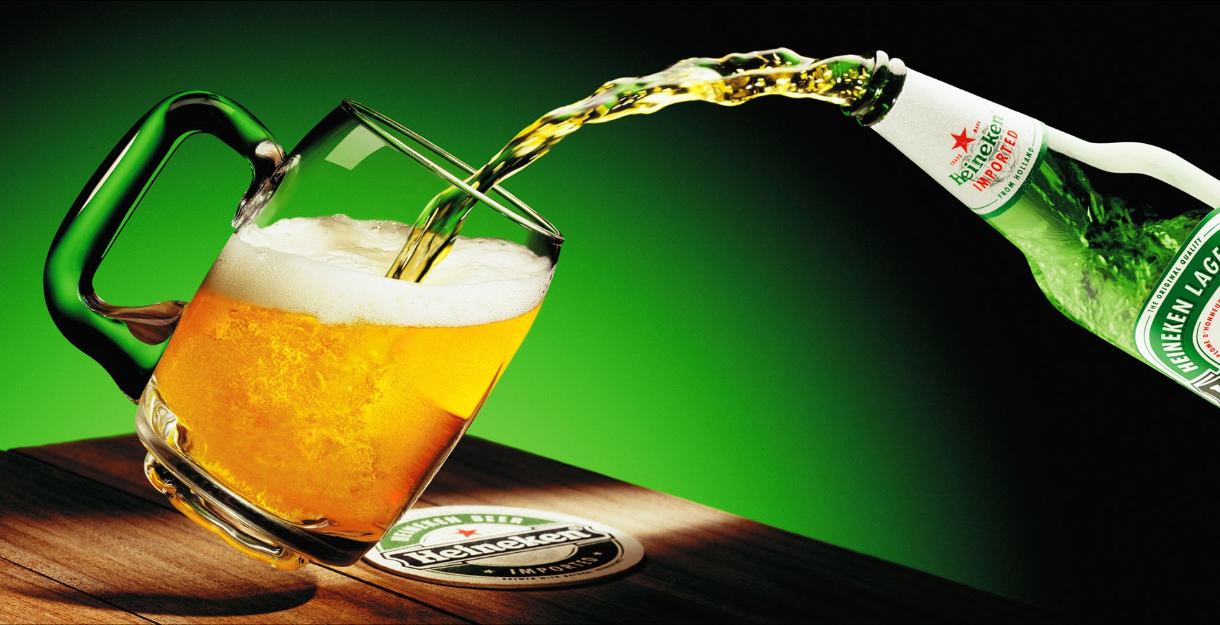 Nalewanie piwa Heineken z butelki do szklanego kufla na drewnianym stole z podkładką pod piwo, zielone tło.