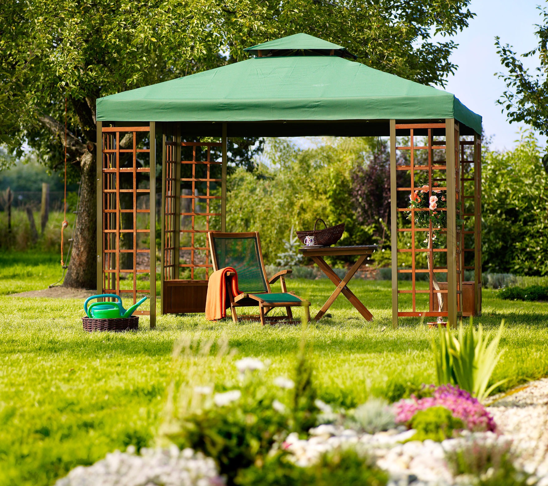 Zadaszona altana ogrodowa z drewnianą konstrukcją, zielonym dachem, rattanowym fotelem z pomarańczowym pledem, stolikiem, kwiatami w donicy i konewką na trawie.