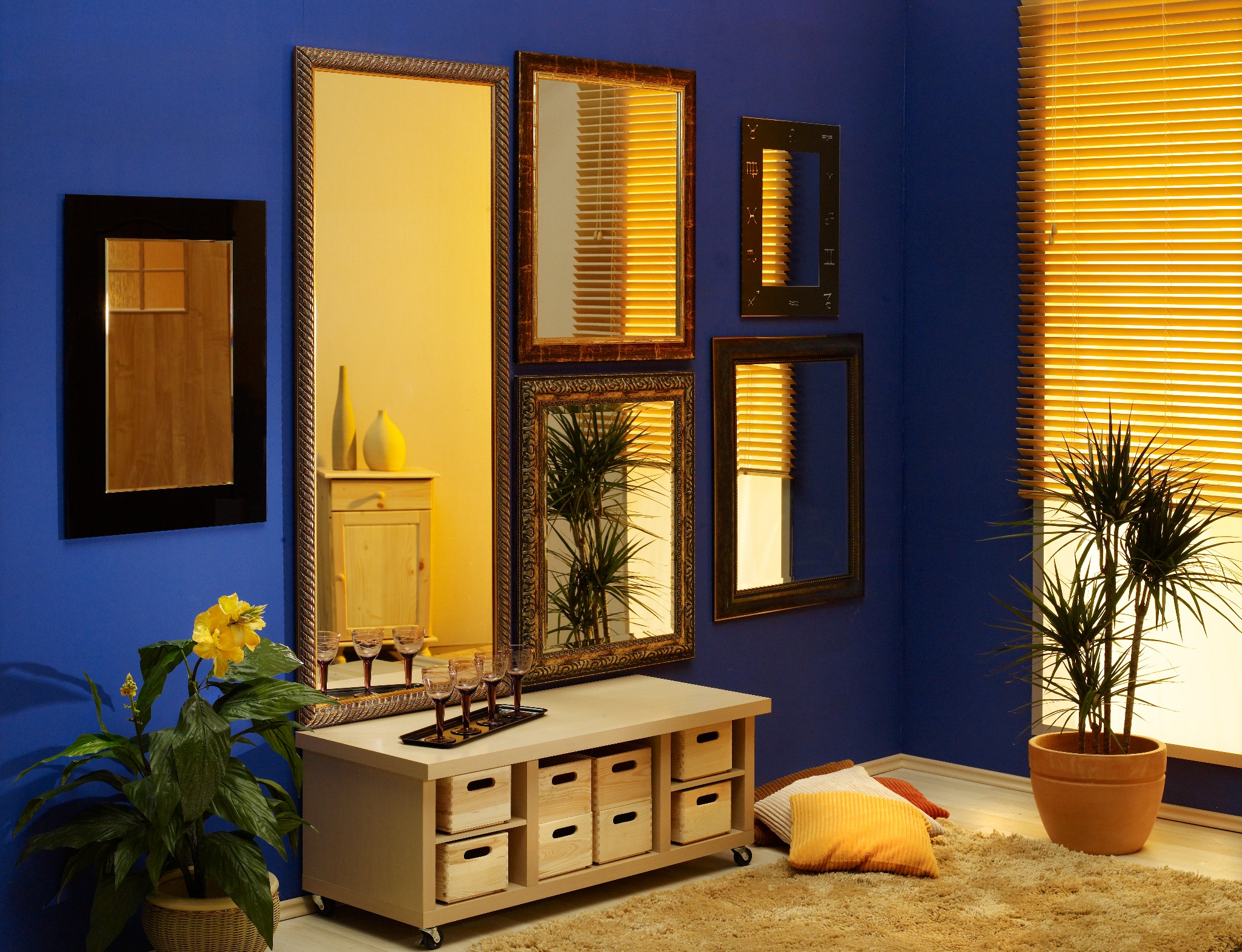 Niebieski pokój z galerią luster w różnych ramach, drewnianą komodą na kółkach z szufladami, kwiatem w koszu i palmą w doniczce, dopełniony dywanem i poduszkami w ciepłych barwach.