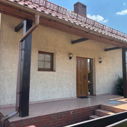 Czystybruk com - Front domu z beżową elewacją, brązowymi rynnami i rurami spustowymi. Widoczny taras z terakoty, drewniane drzwi, okna oraz dachówka ceramiczna z zabezpieczeniami przeciwśniegowymi.