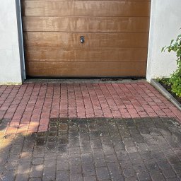 Czystybruk com - Czerwona kostka brukowa przed garażem, częściowo wyczyszczona z brudu i mchu, z wyraźną linią podziału między czystą a zabrudzoną powierzchnią.