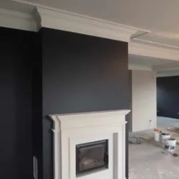 Pokój z czarnymi ścianami i białym, klasycznym kominkiem z metalowym wkładem. Widoczne ślady malowania na posadzce, puszki po farbie w tle. Górna część ściany wykończona dekoracyjnym gzymsem.