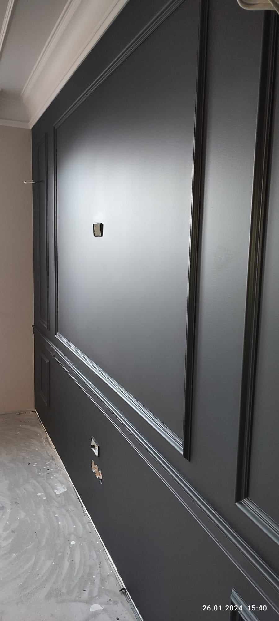 Świeżo pomalowana, ciemnoszara ściana z dekoracyjnymi listwami i gzymsami. Widoczne gniazdka elektryczne i włącznik światła. Podłoga zabezpieczona folią malarską.