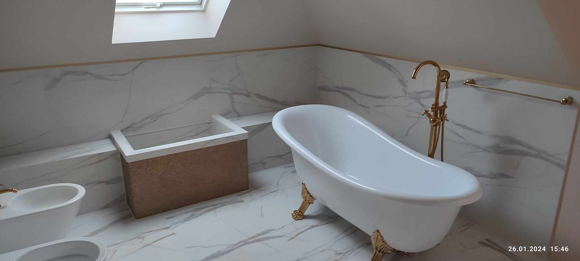 Wykończona łazienka na poddaszu z białą wanną na złotych nóżkach, złotą armaturą, marmurowymi płytkami i oknem dachowym.