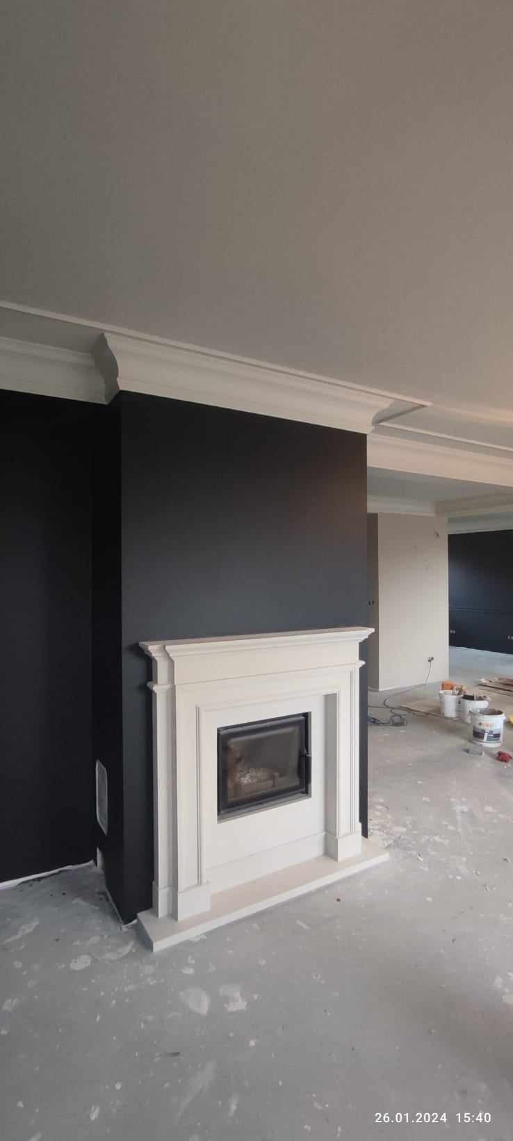 Pokój z czarnymi ścianami i białym, klasycznym kominkiem z metalowym wkładem. Widoczne ślady malowania na posadzce, puszki po farbie w tle. Górna część ściany wykończona dekoracyjnym gzymsem.