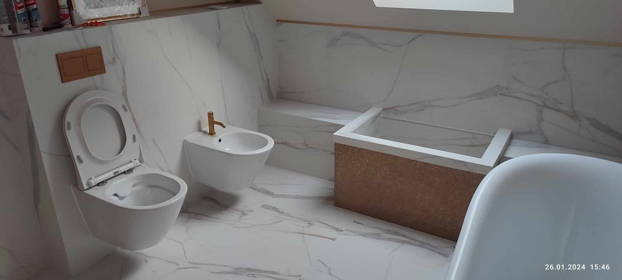 Nowoczesna łazienka z białymi płytkami imitującymi marmur, wisząca toaleta z deską podniesioną do góry, bidet z złotą baterią i zabudowana wanna z brązowym panelem dekoracyjnym.