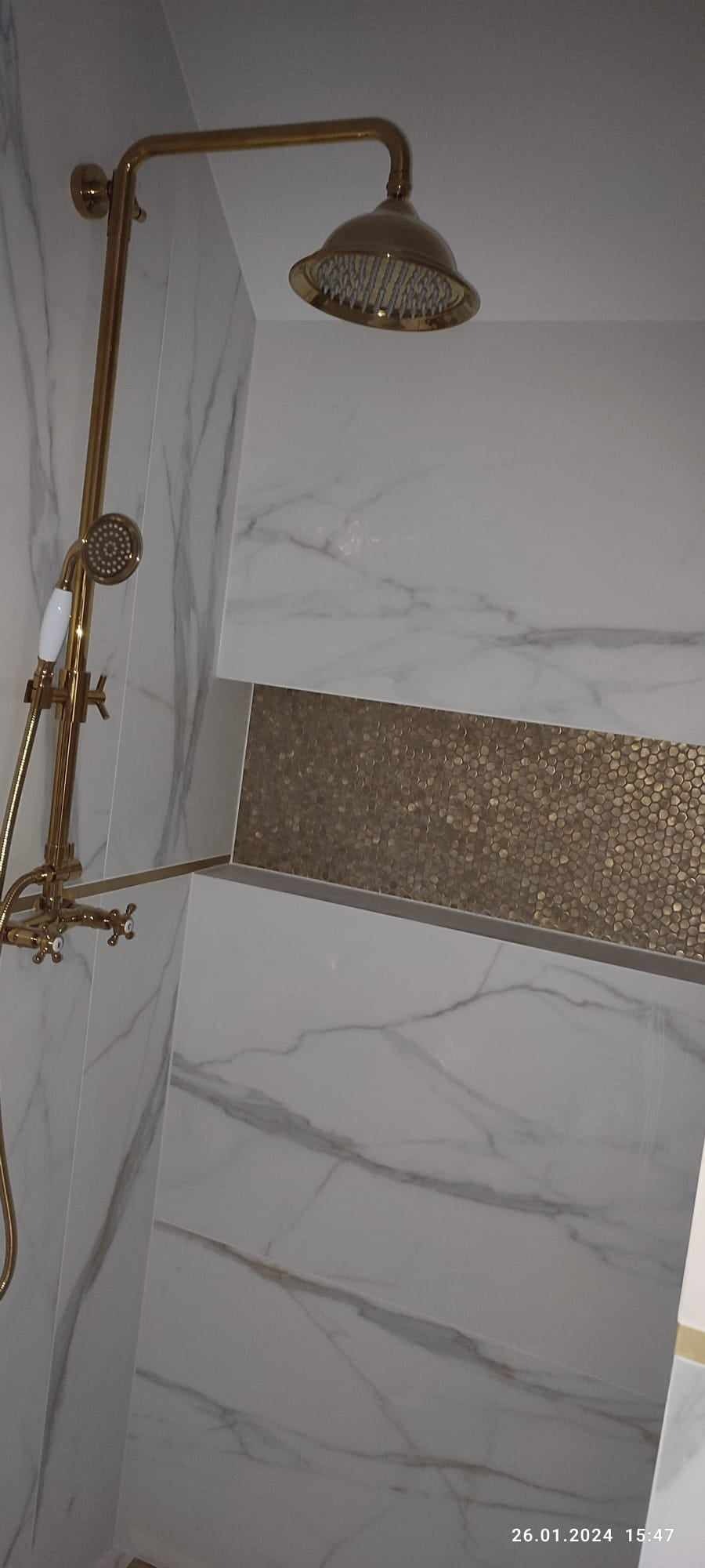 Wykończona łazienka z marmurowymi płytkami i złotą armaturą prysznicową, w tym deszczownicą i słuchawką, z dekoracyjnym pasem mozaiki w odcieniach złota.