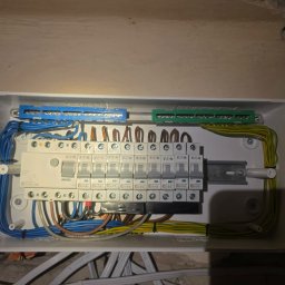 Elektro-Pająk - Biała skrzynka rozdzielcza z rzędem wyłączników nadprądowych Eaton, starannie podłączonymi niebieskimi, brązowymi i żółto-zielonymi przewodami. Widoczne szyny zbiorcze i estetyczne ułożenie...
