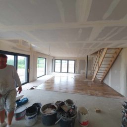 Kajbud - Przestronne wnętrze domu w trakcie prac wykończeniowych: ściany i sufity z płyt gipsowo-kartonowych, częściowo ułożona drewniana podłoga, pracownik w roboczym ubraniu.