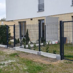 WILCZYŃSKI OGRODZENIA - Nowoczesne, grafitowe ogrodzenie panelowe z bramką, osadzone na betonowej podmurówce, otaczające posesję z budynkiem o jasnej elewacji i zielenią w tle.
