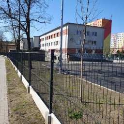 WILCZYŃSKI OGRODZENIA - Nowoczesne, czarne ogrodzenie panelowe z podmurówką betonową, oddzielające chodnik od terenu z budynkami w tle, w słoneczny dzień. Widoczne drzewa i zaparkowane samochody.