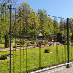 WILCZYŃSKI OGRODZENIA - Nowoczesne ogrodzenie panelowe w kolorze grafitowym, otaczające zadbany ogród z zielonym trawnikiem i kwitnącymi krzewami w słoneczny dzień. Widok z bliska na przęsło.