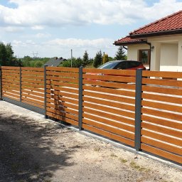 WILCZYŃSKI OGRODZENIA - Nowoczesne ogrodzenie posesji z poziomych desek w kolorze drewna, wsparte na szarych słupkach. W tle dom z czerwoną dachówką i podwójnym garażem.