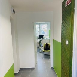 Michał Ożański - Korytarz w odcieniach bieli i zieleni prowadzący do gabinetu dentystycznego z widocznym fotelem i sprzętem. Ściany pomalowane do połowy, szara podłoga z płytek.
