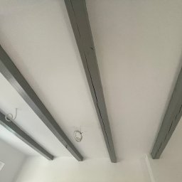 Michał Ożański - Biały sufit z szarymi, pomalowanymi belkami stropowymi. Widoczne kable wystające z sufitu i fragment okna. Ujęcie z dołu, podkreślające geometrię belek.