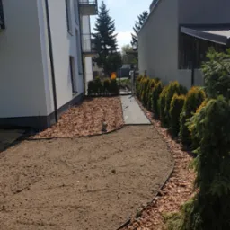 Przygotowany teren pod trawnik z widocznymi obrzeżami, korą ozdobną i ścieżką z płyt betonowych między dwoma budynkami.