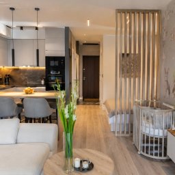 Hana Design Studio - Nowoczesna kawalerka z aneksem kuchennym, drewnianą ażurową ścianką działową i łóżeczkiem dziecięcym. Pastelowe barwy, minimalistyczny design, oświetlenie LED pod szafkami.