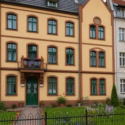 Krzysztof kluczyński - Żółta elewacja kamienicy z zielonymi oknami i drzwiami, ozdobiona ceglanymi detalami. Uroku dodaje balkon z kwiatami i zadbany ogródek z niskim, ozdobnym płotem.