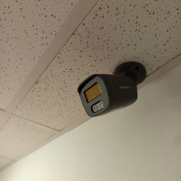 SecureVision - Szara kamera Easycam zamontowana na suficie kasetonowym w pomieszczeniu. Widoczny fragment ściany. Ujęcie z dołu, podkreślające instalację.