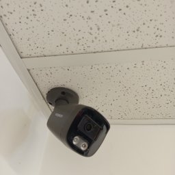 SecureVision - Szara kamera monitoringu marki Ezviz zamontowana pod białym sufitem kasetonowym. Widok z dołu, lekko z boku. Widoczne śruby mocujące i obiektyw kamery.