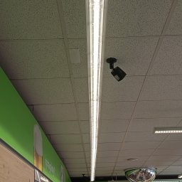 SecureVision - Kamera monitoringu zamontowana na suficie podwieszanym w sklepie, obok oświetlenia liniowego. Widoczny fragment reklamy z motywem piwa i pizzy.