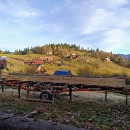 TartakKonopki - Mobilny tartak Wood-Mizer tnie kłodę na tle malowniczego, pagórkowatego krajobrazu z zabudowaniami. Widoczne świeże trociny i fragment pnia na pierwszym planie.