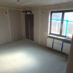 DRYWALL Kamil Nyga - Pokój w trakcie prac wykończeniowych: ściany przygotowane pod malowanie, okno zabezpieczone folią, drabina oparta o ścianę. Widoczne ślady po szpachlowaniu.