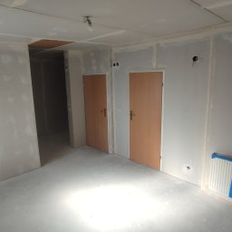 DRYWALL Kamil Nyga - Wnętrze po montażu płyt gipsowo-kartonowych w Łące. Widoczne dwie drewniane ościeżnice drzwiowe, grzejnik zabezpieczony folią i drabina malarska.