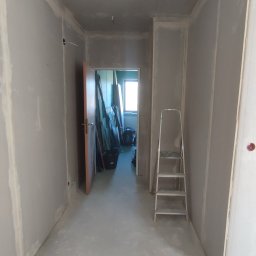 DRYWALL Kamil Nyga - Korytarz w trakcie prac wykończeniowych: ściany z płyt gipsowych, widoczne łączenia, drabina. W tle otwarte drzwi z widokiem na okno i materiały budowlane.