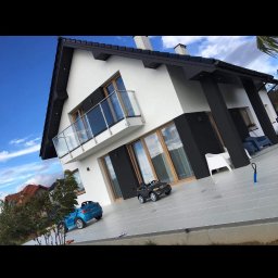 Multi service - Nowoczesny dom z białą elewacją i czarnymi akcentami, balkon z szklaną balustradą, zabawki dziecięce na podjeździe, błękitne niebo w tle.