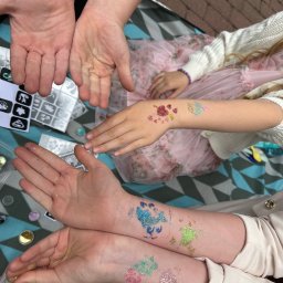 Creative Time - Brokatowe tatuaże na dłoniach dzieci, wykonane podczas imprezy. Widoczne szablony i akcesoria do malowania. Różne wzory i kolory brokatu.