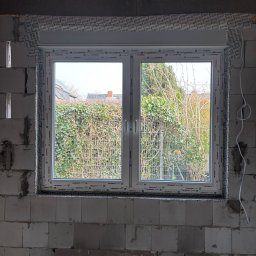 Fenestro - Białe okno PCV Aluplast w surowym murze z betonu komórkowego. Widok na zewnątrz, gdzie widać zieleń i fragment dachu sąsiedniego budynku. Okno z roletą zwiniętą u góry.