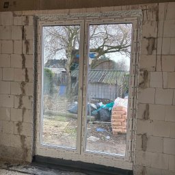 Fenestro - Białe okno PCV zamontowane w surowym murze z betonu komórkowego. Widok na zewnątrz: drzewo, ogród, worki z cementem. Folia ochronna na ramie okiennej.