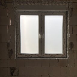 Fenestro - Białe okno PCV Aluplast zamontowane w surowej ścianie z betonu komórkowego. Widoczne uszczelnienia i taśmy izolacyjne. Etap montażu okna w nowym budynku.