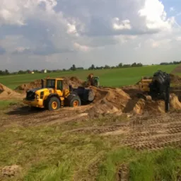 Żółte ładowarki Volvo pracują na polu, usypując kopce ziemi. W tle zielone pola i pochmurne niebo. Widoczne ślady opon na ziemi.
