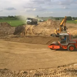 Walec w trakcie utwardzania terenu pod budowę na rozległym placu, w tle koparka i ciężarówka. Widoczne ślady opon na ziemi i zielone pola w oddali.