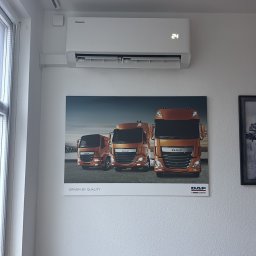 Klima Neketech - Biały klimatyzator Hisense na ścianie biura, pod nim plakat z trzema pomarańczowymi ciężarówkami DAF. Widoczne okno i fragment lampy sufitowej.