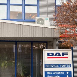 Klima Neketech - Biała jednostka klimatyzacji Hisense zamontowana na szarym budynku biurowym z oknami, w których widać sylwetkę osoby. Na pierwszym planie znak DAF Service Parts.