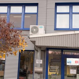 Klima Neketech - Biała jednostka zewnętrzna klimatyzacji Hisense zamontowana na elewacji budynku biurowego z niebieskimi ramami okiennymi i metalową fasadą w jesiennej scenerii.