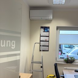 Klima Neketech - Biały klimatyzator Mitsubishi zamontowany w biurze, widoczna drabina, kalendarz i odkurzacz. Ujęcie z perspektywy podłogi, wnętrze biurowe z oknem.