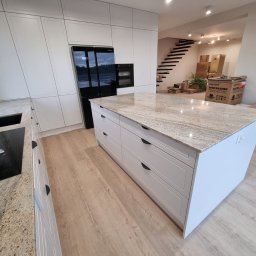 KNK Meble - Biała kuchnia z wyspą i blatem z kamienia. W tle schody i kartony. Nowoczesny design, minimalistyczne fronty szafek, czarny sprzęt AGD. Jasne drewno na podłodze.