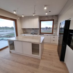 KNK Meble - Jasna kuchnia na wymiar z wyspą i blatem z kamienia. Nowoczesne oświetlenie punktowe. Duże okno z widokiem na zewnątrz. Minimalistyczny design.