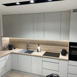 KNK Meble - Nowoczesna kuchnia z szarymi szafkami, drewnianym blatem i oświetleniem LED. Widoczny zlew, płyta indukcyjna i piekarnik. Minimalistyczny design.
