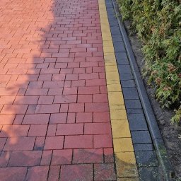 Czysty bruk - Czerwona kostka brukowa z żółtym i szarym obrzeżem, prowadząca do metalowej bramy, obok żywopłotu. Widoczne cienie drzew na kostce.