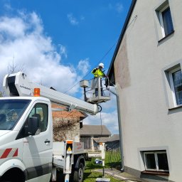 Czysty bruk - Mycie elewacji domu z podnośnika koszowego w Tomaszowie Lubelskim. Pracownik w kasku i kamizelce czyści ścianę budynku z zabrudzeń. Widoczne efekty czyszczenia.