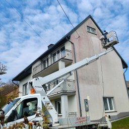 Czysty bruk - Pracownik na podnośniku koszowym czyści elewację dwupiętrowego domu z balkonami. Widoczny fragment elewacji przed i po czyszczeniu. Błękitne niebo w tle.
