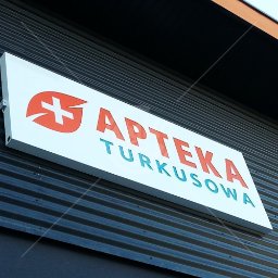 Kiwi Gifts Drukarnia Reklama - Biały szyld apteki z czerwonym logo i napisem 'Apteka Turkusowa' na tle szarej ściany z blachy falistej.