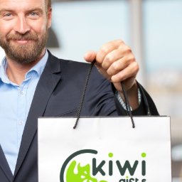 Kiwi Gifts Drukarnia Reklama - Uśmiechnięty mężczyzna w garniturze prezentuje białą torbę z logo Kiwi Gifts, widoczne biurowe tło.