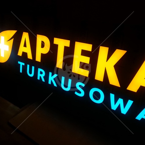 Podświetlany szyld apteki z żółtymi literami i niebieskim napisem 'Turkusowa' na czarnym tle.